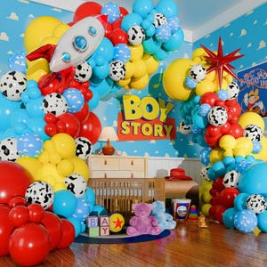 May include: A colorful Toy Story-themed party scene with balloons, a rocket, and the words "BOY STORY." The room features a crib, teddy bears, and wooden blocks spelling "BAY." Balloons are red, yellow, blue, and white.