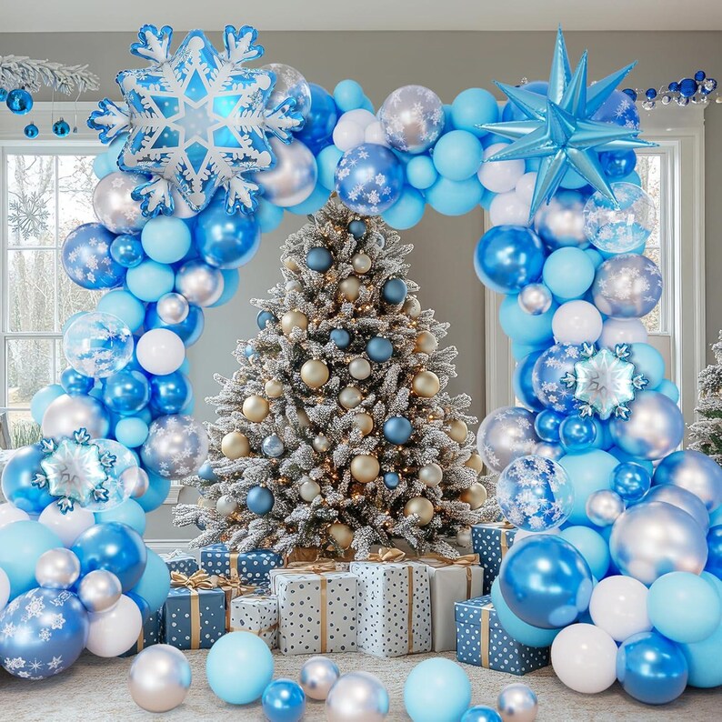 May include: A decorated Christmas tree with gold and blue ornaments, surrounded by a balloon arch in shades of blue, silver, and white. Snowflake and star-shaped balloons add to the festive decor. Gift boxes are placed at the base of the tree.