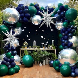 May include: A decorative balloon arch featuring a mix of green, navy blue, and silver balloons. The arch is adorned with star-shaped decorations and is set against a dark background with star cutouts. The scene includes a table with plants.