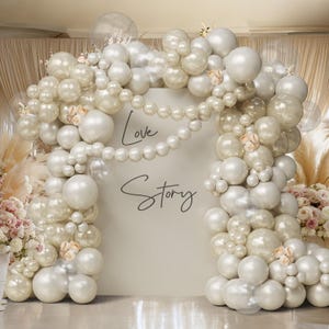 May include: A decorative backdrop for a wedding or event, featuring a cream-colored panel with the words "Love Story" in elegant script. The panel is framed by a large arrangement of white and pearl balloons, with a string of pearls and small floral accents.