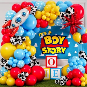 May include: A colorful balloon arch featuring red, blue, yellow, and cow-print balloons. A rocket-shaped balloon and a red star-shaped balloon are included. The backdrop reads "It's a Boy Story" with block letters and toy blocks.
