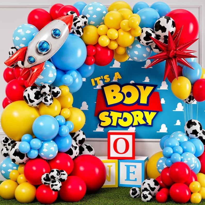 May include: A colorful balloon arch featuring red, blue, yellow, and cow-print balloons. A rocket-shaped balloon and a red star-shaped balloon are included. The backdrop reads "It's a Boy Story" with block letters and toy blocks.