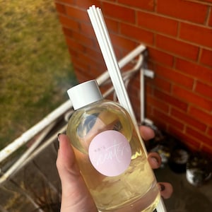 May include: A clear glass reed diffuser bottle filled with a light yellow liquid and containing white flower petals. The bottle has a white cap and a round pink label with the text KB'S scents in white script. Several white reed sticks are included.