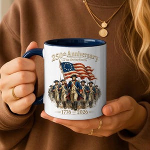 May include: A white ceramic mug with a dark blue handle and interior. The front features a graphic of Revolutionary War soldiers marching under an American flag with the text 250th Anniversary of the United States and 1776-2026 printed in gold.