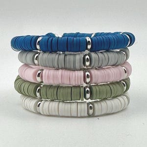 May include: Five stacked bracelets in a variety of colours. Each bracelet features flat, round beads in blue, grey, pink, green, and white. Silver-coloured metal spacers are between the beads. The bracelets are stacked together.