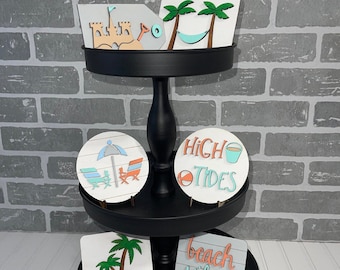 May include: A three-tier black tiered stand with five wooden signs featuring beach-themed designs. The signs include a sandcastle, palm trees, an umbrella, a beach chair, a beach ball, and the text "High Tides" and "Beach Vibes".