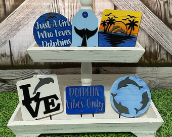 May include: A two-tiered white wooden tiered tray with five wooden signs featuring dolphin designs. The signs say "Just a Girl Who Loves Dolphins", "Dolphin Vibes Only", "Love", and a sign with a sunset and palm trees.