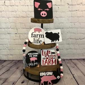 May include: A tiered wooden stand with black metal accents displays various farmhouse-themed signs featuring pink and black pigs. The signs include phrases like "farm life", "welcome to our pig pen", and "life is better on the farm".