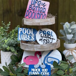 May include: A three-tiered wooden tray display with various summer-themed wooden signs. The top tier features a pink and blue sign that reads "Life is Better Poolside". The middle tier has a blue sign that reads "Pool Vibes" and a white sign that reads "Pool Hair Don't Care". The bottom tier has a pink flamingo, a blue and white pair of goggles, and a blue sign that reads "Made for Sunny Days".