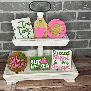 Könnte beinhalten: Ein weißes, zweistöckiges Tablett mit dekorativen Holzschildern zum Thema Tee. Die Schilder zeigen Sätze wie "Tea Time" und "But first tea" mit rosa und grünen Akzenten. Eine Teekanne und eine Teetasse sind ebenfalls abgebildet.