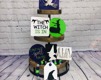 May include: A tiered wooden stand with three shelves decorated with Halloween-themed wooden signs. The top shelf has a sign with a silhouette of a witch flying on a broomstick. The middle shelf has a sign that says "The Witch Is In" and a green sign with a silhouette of a witch. The bottom shelf has a sign that says "I Put A Spell On You" and a white sign with a silhouette of a witch's hat. The stand is decorated with a black and green beaded garland.