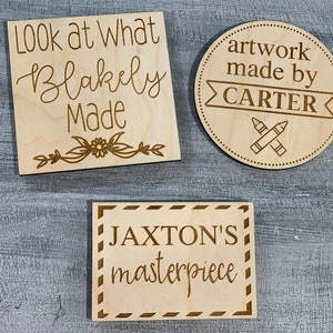 May include: Three wooden signs with personalized messages. The first sign says "Look at what Blakely Made" with a floral design. The second sign says "artwork made by CARTER" with a banner and crossed crayons. The third sign says "JAXTON'S masterpiece" with a decorative border.