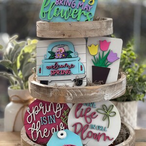 May include: A tiered wooden tray display featuring spring-themed wooden signs. The top sign reads "April showers bring May flowers". The middle tier has a sign with a truck that says "Welcome Spring" and a sign with three yellow, green, and purple tulips in a pot. The bottom tier has a sign that says "Spring is in the air" and a sign that says "Live life in full bloom".