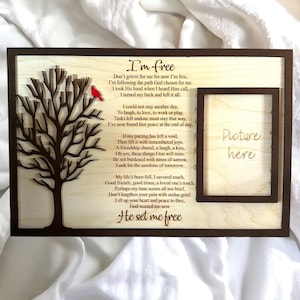 May include: Wooden memorial picture frame with a laser-cut tree and a red bird. The frame includes a poem titled "I'm Free" and a space to insert a picture. The frame is made of light-colored wood with a dark brown border.