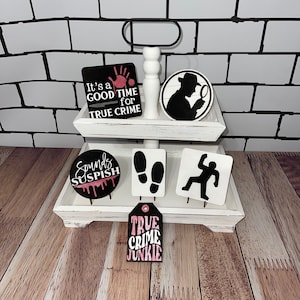 May include: A two-tiered white wooden tray with black and white true crime themed decorations. The decorations include a handprint, footprints, a detective, and the phrases "It's a good time for true crime" and "True Crime Junkie."