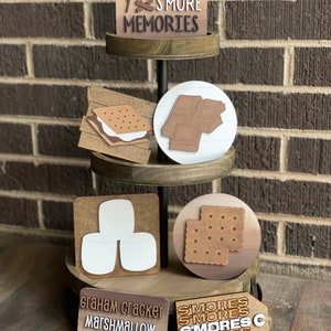 以下が含まれることがあります： 「Making S'more Memories」と書かれた看板が付いた、3段のウッドスタンド。スタンドは、グラハムクラッカー、マシュマロ、チョコレートバー、スモアのウッドカットアウトで飾られています。スタンドには、「Graham Cracker, Marshmallow, Chocolate」と書かれた看板と、「S'mores」が複数回書かれた看板も付いています。