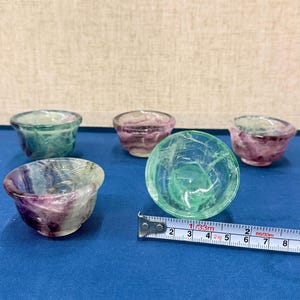 以下が含まれることがあります： 緑と紫の色合いの4つの小さな半透明のボウルが、青い表面に配置されています。メジャーテープは、ボウルの直径が約5cmであることを示しています。ボウルはユニークで自然な石の外観をしています。