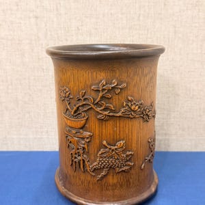 May include: A cylindrical wooden container with intricate carvings. The container features detailed relief carvings of floral and foliate designs, including a grape cluster. The wood has a warm, brown tone.