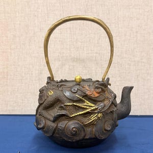 以下が含まれることがあります： 龍のモチーフが付いた、装飾的な鋳鉄製のティーポット。 ティーポットは真鍮製のハンドルとアクセント、注ぎ口が特徴です。 龍のデザインは、金とブロンズの色合いで詳細に描かれています。 ティーポットの高さは約15cmです。