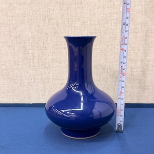 以下が含まれることがあります： 丸みを帯びたボディと細い首を持つコバルトブルーのセラミック花瓶。 花瓶の高さは約18cmです。 花瓶の横に巻尺が置かれています。