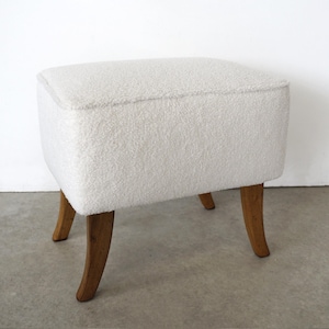 Può includere: Un piccolo pouf bianco imbottito di forma rettangolare. Il pouf ha una superficie morbida e strutturata ed è sostenuto da quattro gambe in legno curve. Le gambe sono di colore marrone caldo, in contrasto con la parte superiore bianca.