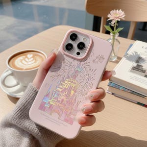 May include: A pink phone case featuring a castle design with fireworks. The case has a light pink border around the camera lenses. The text at the bottom of the case reads: "The happiness of a moment of freedom with the joy you bring."