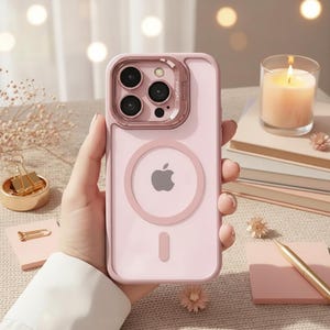 May include: A pink iPhone case with a clear back and a pink frame. The case has a circular MagSafe charging ring with the Apple logo and a camera cutout. The case is held in a person's hand.