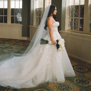 May include: A white bridal gown with floral detailing and a long train. The bride carries a bouquet of white roses. A long veil is draped down the back. The setting is a room with large windows.