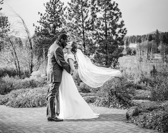 May include: A bride and groom kiss in a black and white photo. The bride's veil is blowing in the wind. They are standing on a brick path in front of a forest.