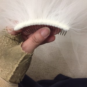 May include: White bridal veil with a comb attached to the edge. The comb is made of metal and has a white fabric covering.
