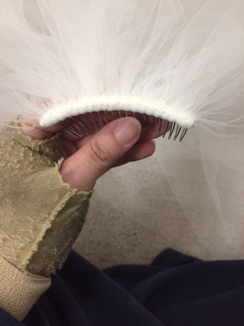 May include: White bridal veil with a comb attached to the edge. The comb is made of metal and has a white fabric covering.