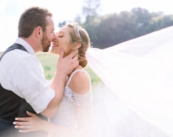 May include: A couple embraces outdoors, the woman wearing a white wedding dress with a beaded bodice and a tiara. A long, flowing veil billows in the wind. The man wears a white shirt, black vest, and tie. The scene is bathed in soft sunlight.