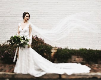 May include: A bride in a white wedding dress with a long train and lace sleeves stands in front of a white brick wall. She holds a bouquet of white and green flowers. A long veil flows in the wind.