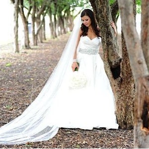 May include: A bride in a white wedding dress with a long veil stands in a grove of trees. She is holding a bouquet of white flowers.