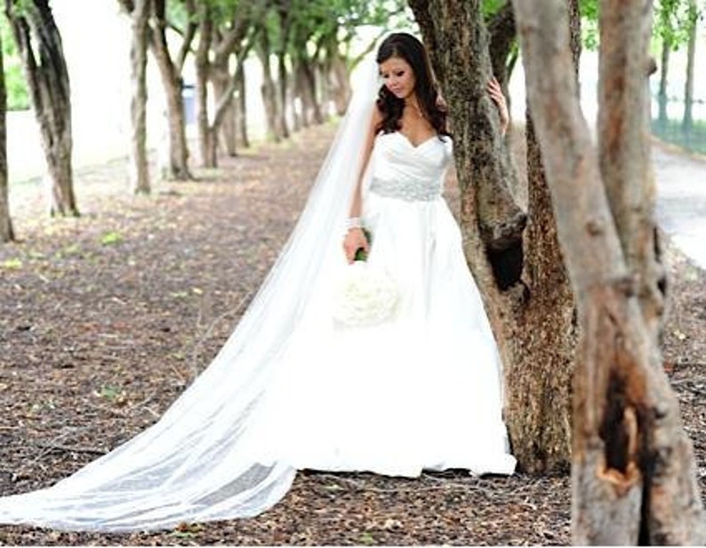 May include: A bride in a white wedding dress with a long veil stands in a grove of trees. She is holding a bouquet of white flowers.