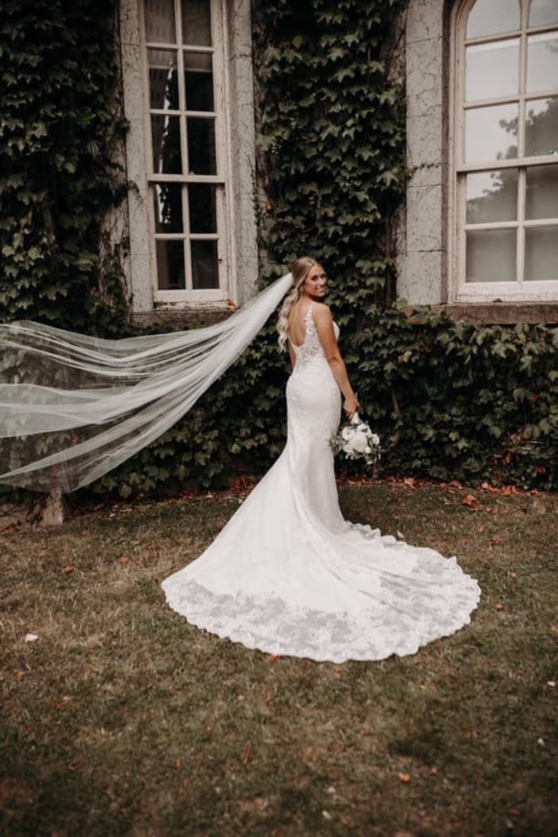 May include: A bride in a white lace wedding dress with a long veil, standing in front of a building covered in ivy. She is holding a bouquet of white flowers.