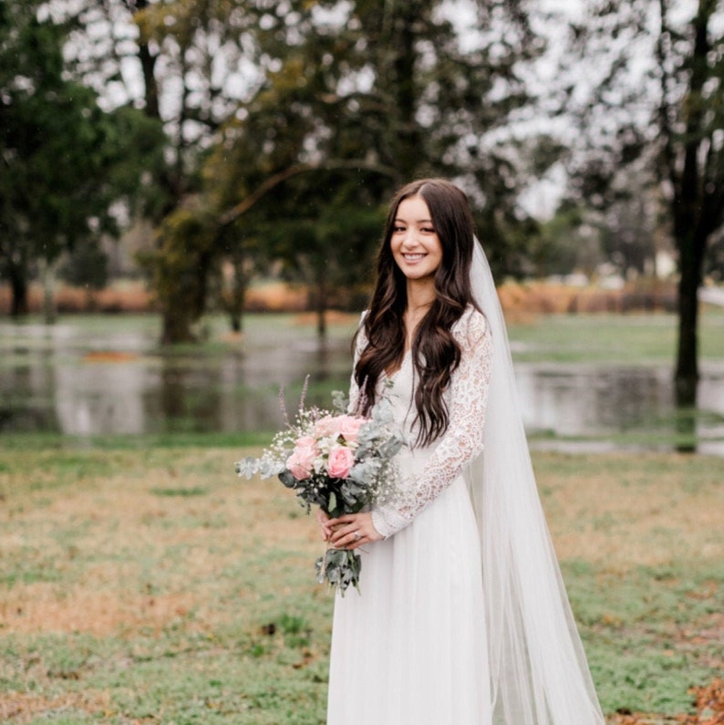 May include: A bride in a white lace dress and veil holds a bouquet of pink and white flowers. She is standing in a grassy field with trees in the background.