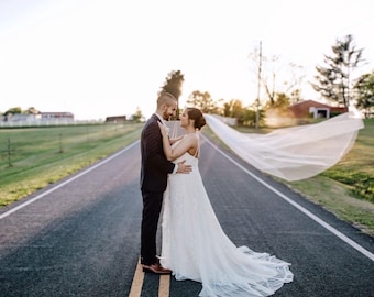 May include: A bride and groom stand on a paved road, embracing and looking at each other. The bride is wearing a white wedding dress with a long veil that is flowing in the wind. The groom is wearing a black suit and tie. The road is lined with trees and grass.