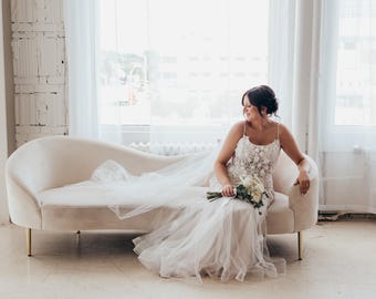 May include: A bride in a white wedding dress with floral detailing sits on a cream-colored curved sofa with gold legs. She holds a bouquet of white flowers. The dress has a long, flowing train. The room has large windows and white brick.