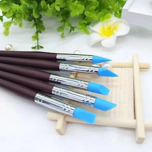 May include: A set of six brown-handled art tools with silver ferrules and blue silicone tips. The tips have various shapes, including pointed and angled designs. The tools are arranged on a light wooden structure, with green foliage and a white flower in the background.