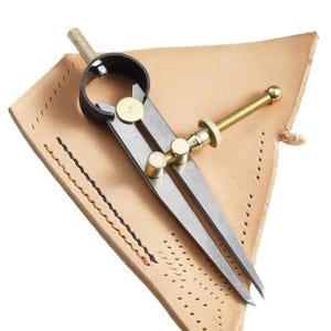 May include: A metal compass with brass accents, resting on a tan leather sheath. The compass has a black top and a threaded adjustment screw. The leather sheath has decorative stitching and a triangular shape.