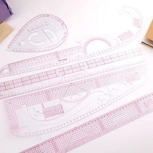 May include: A selection of transparent, pink-tinted plastic rulers and templates for sewing and design. The set includes various shapes, such as straight rulers, curved templates, and a teardrop-shaped template with a design. The rulers are marked with centimetre measurements.