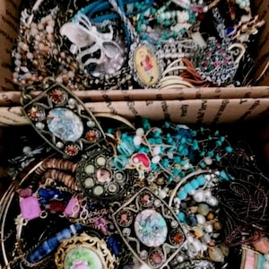 Può includere: Una vasta collezione di bigiotteria assortita contenuta in una scatola di cartone, composta da varie collane, bracciali, spille e anelli con perline colorate, dettagli metallici, motivi floreali e pietre decorative di forme diverse.