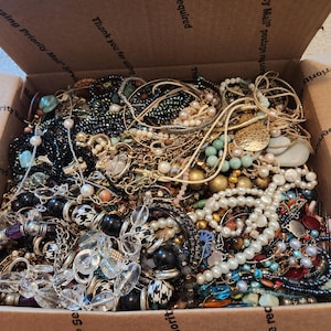 Può includere: Una grande scatola di cartone riempita con una collezione varia di bigiotteria, tra cui varie collane, bracciali e orecchini con perle, cristalli trasparenti, perline nere, catene color oro e perline sferiche decorate.