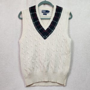 May include: Cream-colored cable knit sweater vest with a V-neck. The neckline is trimmed with a plaid pattern in navy, red, and green. The Polo by Ralph Lauren label is visible at the top.