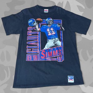 May include: A navy blue t-shirt featuring a graphic of a football player in a blue uniform throwing a football. The shirt has the words "GIANTS" and "NEW YORK" in a blue and pink design, along with the name "PHIL SIMMS" and the number "11".