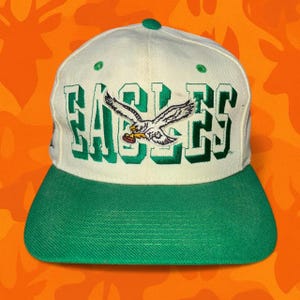 May include: A cream-colored baseball cap with a green brim. The word "EAGLES" is embroidered in green across the front, with an eagle logo in the center. The cap has a green button on top.