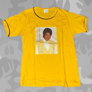 May include: Yellow t-shirt featuring a portrait of Michael Jackson. The shirt has a black trim around the collar and sleeves. The portrait shows Michael Jackson wearing a yellow vest and white shirt.