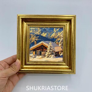 Pode incluir: Uma pequena pintura quadrada numa moldura dourada decorativa que retrata uma cabana de madeira coberta de neve numa paisagem montanhosa com um pinheiro em primeiro plano. O texto SHUKRIASTORE está impresso a branco na parte inferior.