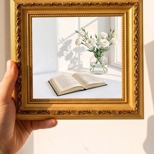 Könnte beinhalten: Ein quadratischer, verzierter goldfarbener Bilderrahmen mit einer Illustration eines aufgeschlagenen Buches und einer klaren Glasvase mit weißen Blumen. Das Kunstwerk zeigt eine sonnendurchflutete Szene mit weichen Schatten auf einer weißen Fläche.